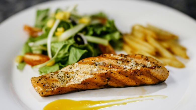 Grilled salmon on a white plate with a small amount of sauce with salad anf ries in the background