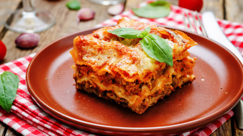Meat lasagna on a red plate sitting on top red and white checkered cloth on wood table.