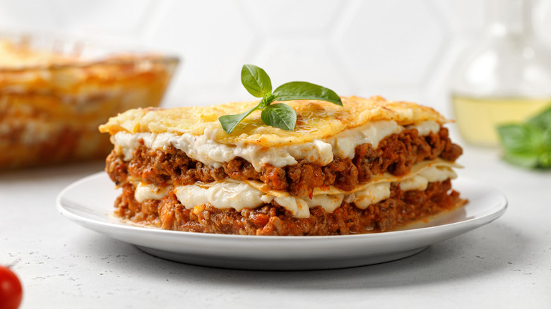 Piece of lasagna with bolognese meat sauce, cheese, and basil on white plate.