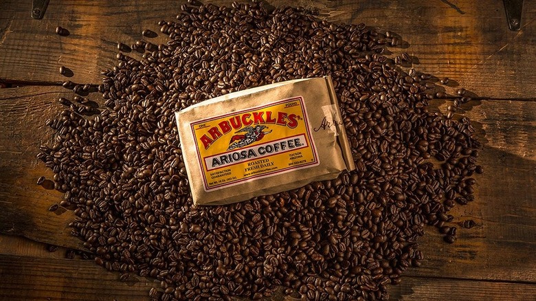 A one-pound bag of Arbuckles' Ariosa Coffee lies atop a bed of coffee beans on a flat wooden surface, which appears to be the top of a storage chest..