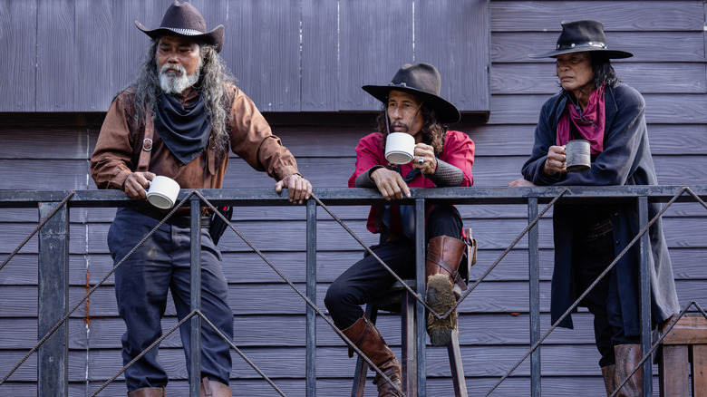 Three older Hispanic cowboys drink coffee out of ceramic and metallic mugs on a deck in the daylight, leaning on a black railing as they look towards the ground below.