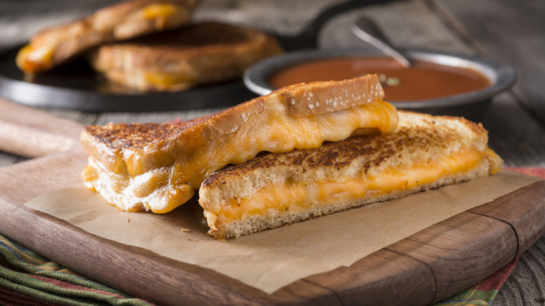A golden grilled cheese sliced in half on the diagonal on a cutting board by a bowl of tomato soup.