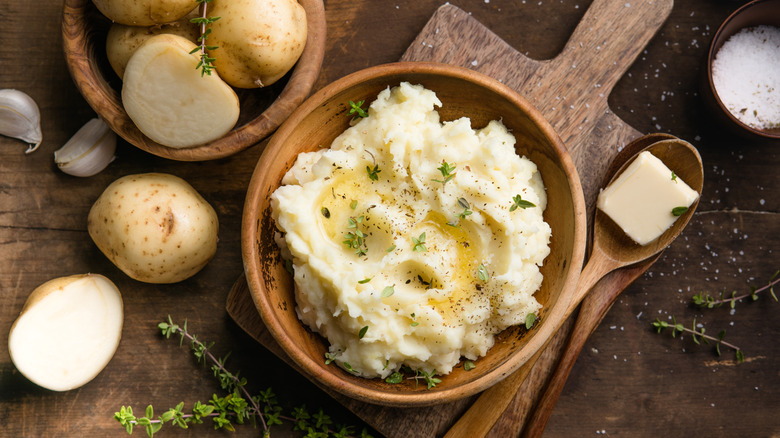 Wooden bowl of mashed potatoes topped with butter and herbs on a cutting board with raw potatoes in the background
