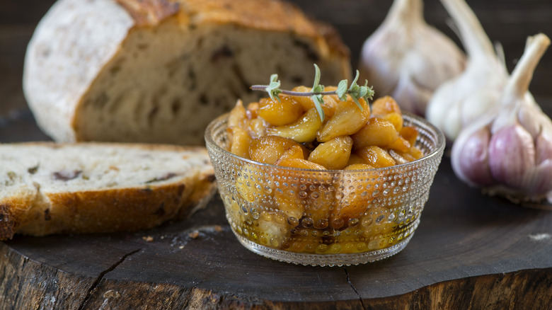 Garlic confit in a clear small bowl with rustic bread and raw garlic in the background.