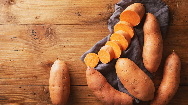Whole sweet potatoes, plus a sliced one, on a wooden table.