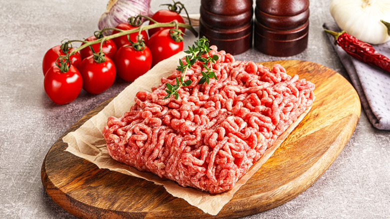 A portion of raw ground beef on a wooden board with herbs and tomatoes.