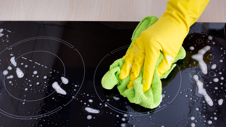 Yellow gloved hand holding green microfiber towel cleaning glass stovetop.