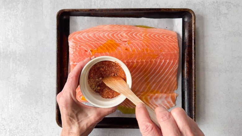 Mixing salmon seasoning spices in small bowl with wooden spoon above salmon filet on baking sheet