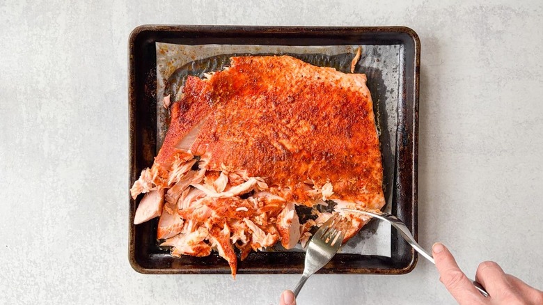 Flaking baked salmon filet into chunks using forks on parchment-lined baking sheet