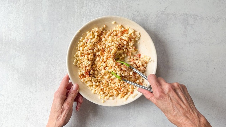 Tossing raw corn kernels with olive oil and spices in shallow bowl with tongs