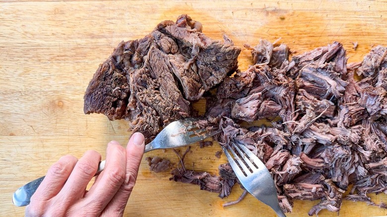 Shredding cooked beef chuck with forks on cutting board