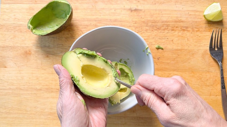 Scooping pitted avocado flesh into bowl with spoon