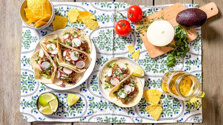 Slow cooker barbacoa tacos on serving platter and plate on table with chips, avocado, onion, limes, and tomatoes
