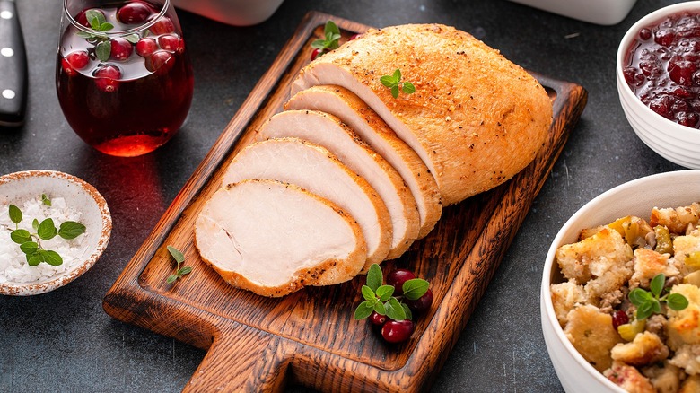 Half-sliced turkey breast on a wooden cutting board surrounded by bowls of stuffing and cranberry sauce