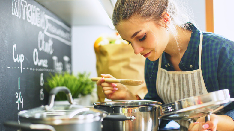 Person holding the lid to a soup pot in one hand and a wooden spoon in the other, looking down into a pot cooking on a stove