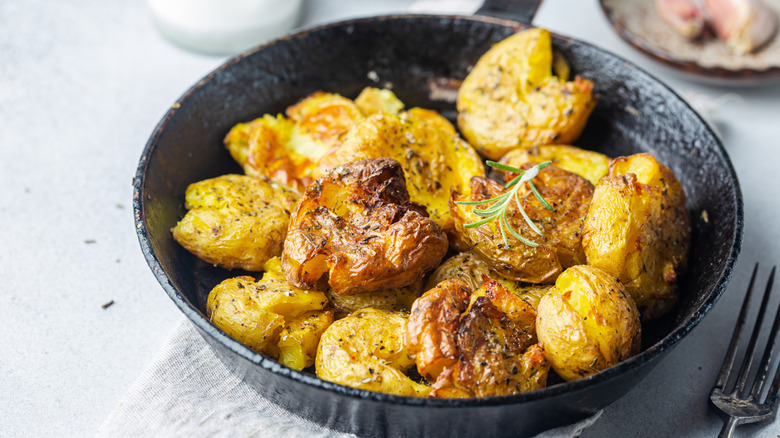 Smashed potatoes in skillet with fresh herb garnish on white table.