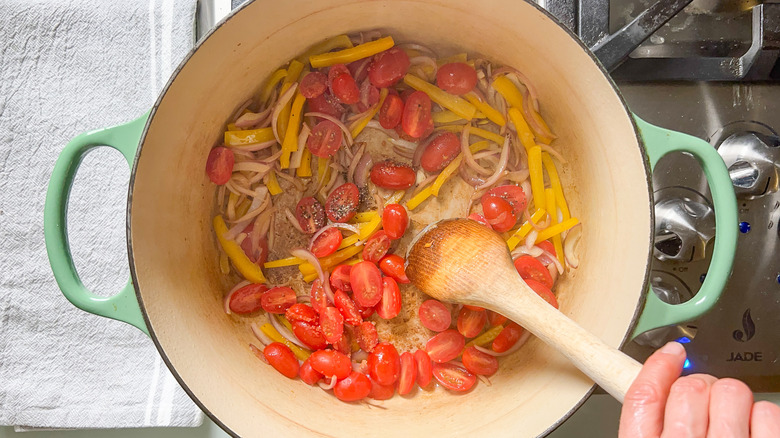 Cooking red onion, yellow bell pepper, grape tomatoes, and garlic in pot with wooden spoon on stove top