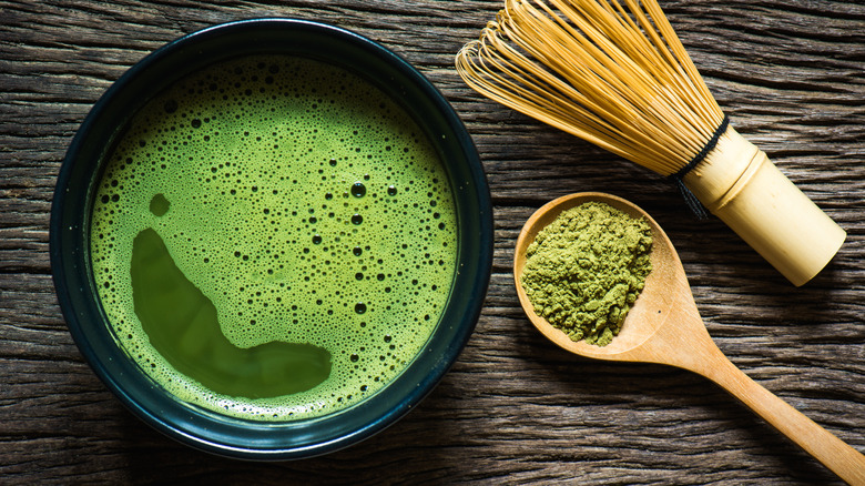 A cup of matcha tea next to a spoon filled with matcha powder and a whisk