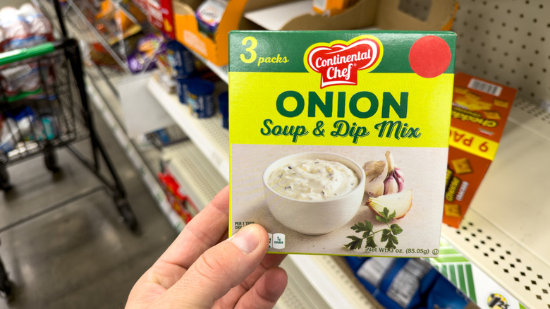 A person's hand is seen holding a box of onion soup mix in a grocery store.