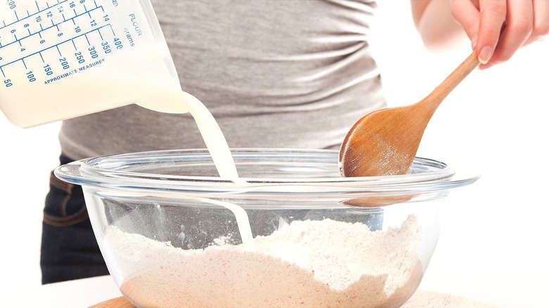 Hand pouring buttermilk from a measuring cup into a clear bowl of flour