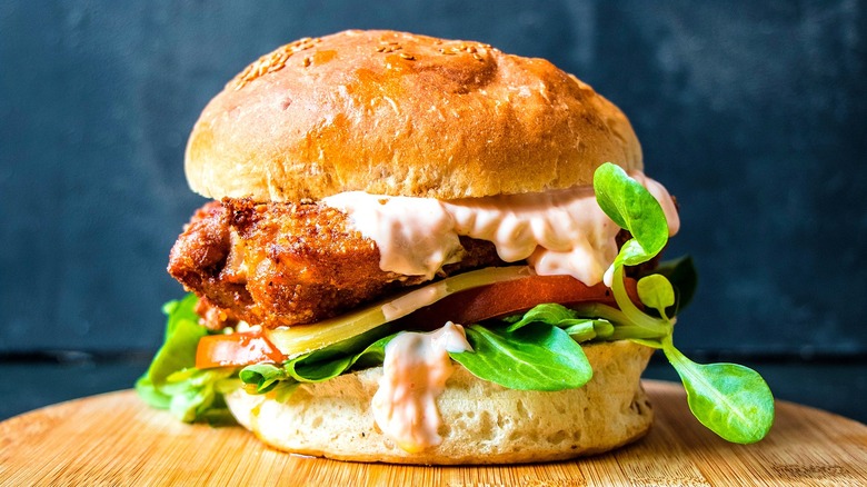 Fried chicken sandwich with fresh greens, sliced pickle, tomato, and sriracha mayo on a round, wooden serving board