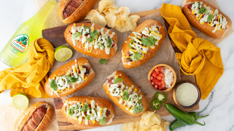 Sonoran hot dogs surrounded by various toppings, napkins, chips, and bottle of Jarritos