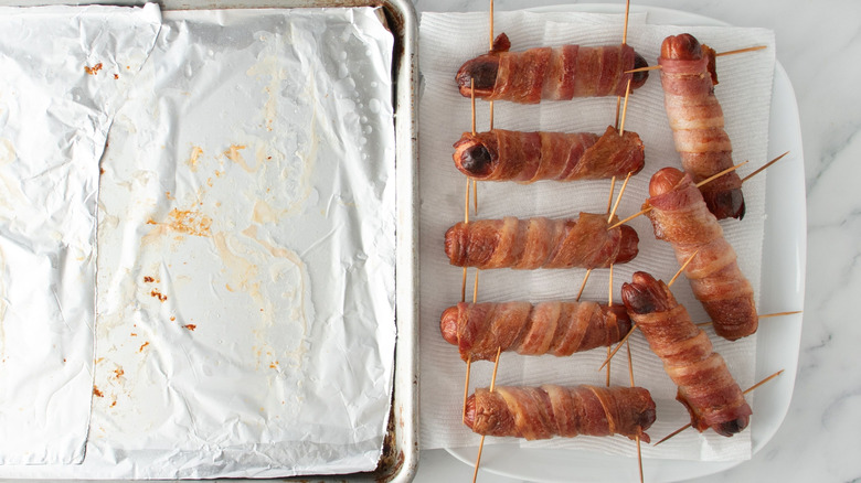 Aluminum-lined baking sheet next to bacon-wrapped hot dogs on plate