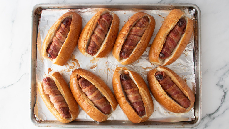 Bacon-wrapped hot dogs in buns on baking sheet