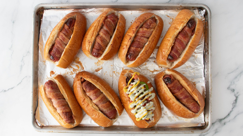 Bacon-wrapped hot dogs on baking sheet with one garnished with sauces