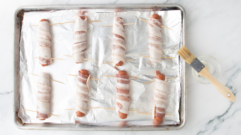 Bacon-wrapped hot dogs on baking sheet next to pastry brush