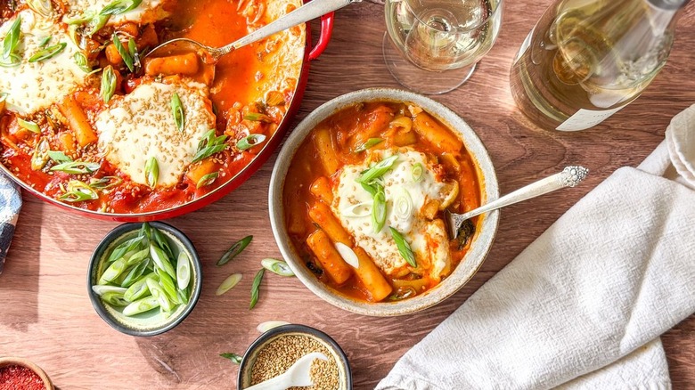 Spicy baked tteokbokki with burrata in pan and serving bowl with garnishes and wine
