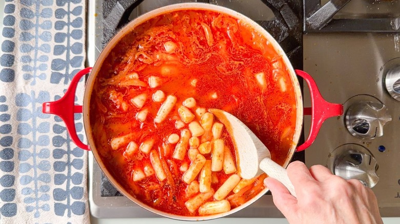 Stirring tteokbokki in red pot on stovetop with wooden spoon