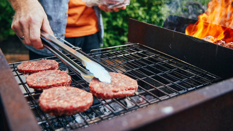 A person's hands are seen using tongs to manage burgers on a grill.