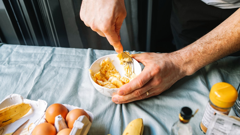 A hand mashing bananas with a fork in a small bowl, surrounded by other  banana bread ingredients on the table