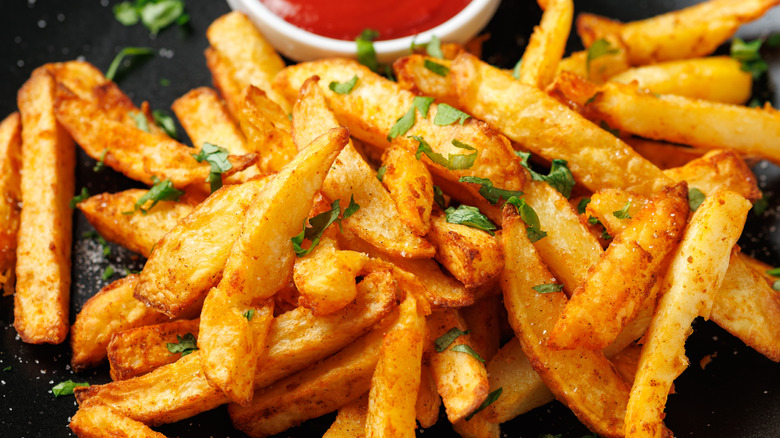 A closeup of French fries sprinkled with herbs and a cup of red sauce in the background