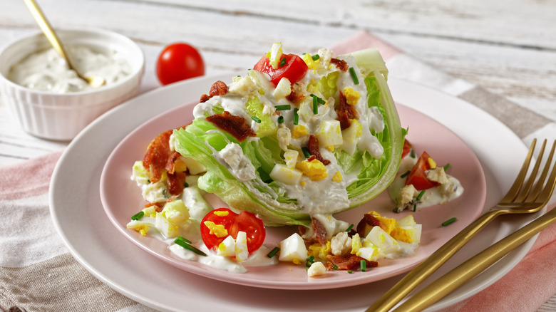 A wedge salad photographed from an elevated angle on a plate with silverware, next to a ramekin of dressing with a spoon and a cherry tomato