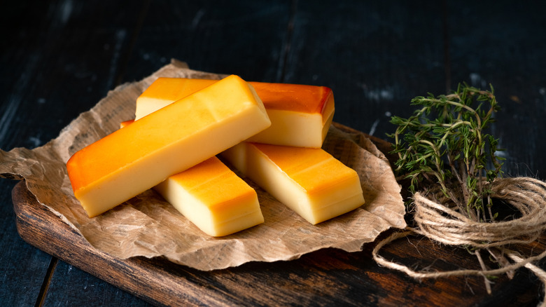 Chunks of smoked cheese on brown paper, resting atop a wooden board, with a sprig of herbs beside them