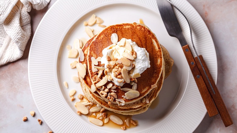 Top-down view of pancakes with whipped cream, maple syrup, and sliced almonds on a white plate