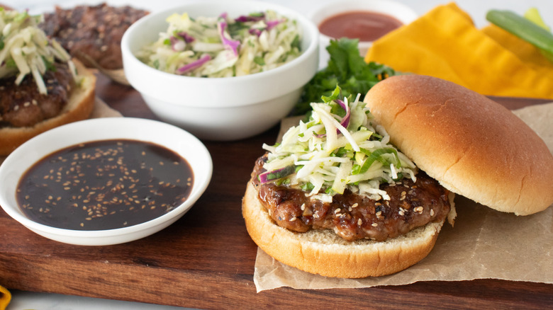 A side shot of an assembled burger topped with slaw surrounded by condiments and toppings