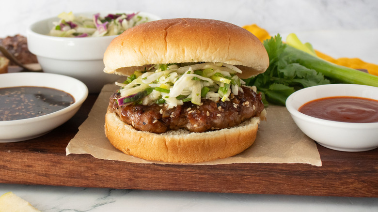 A side shot of an assembled burger topped with slaw surrounded by condiments and toppings