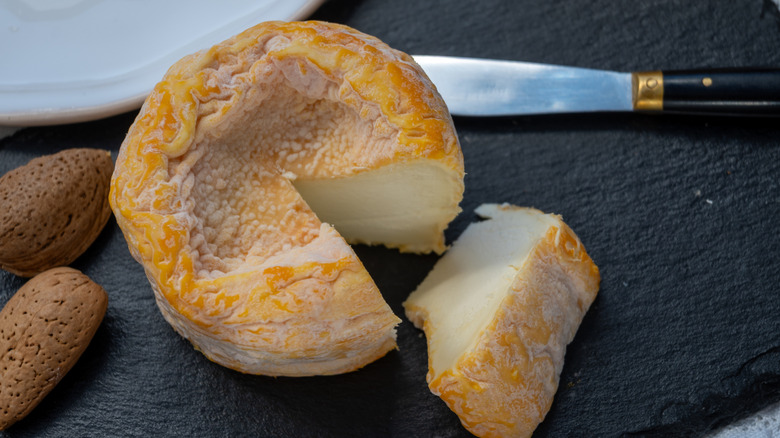 A small wheel of washed-rind cheese with a wedge on a slate next to nuts and a knife