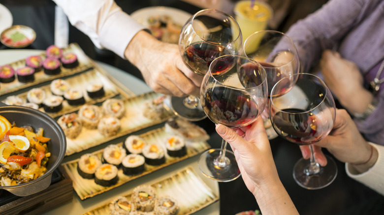 Hands toast with red wine over a table full of sushi.