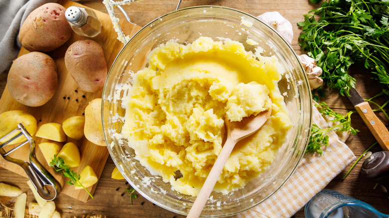 A glass bowl of mashed potatoes with a wooden spoon in it, mid-preparation, surrounded by whole and cut boiled potatoes, a knife and peeler, salt, and parsley.