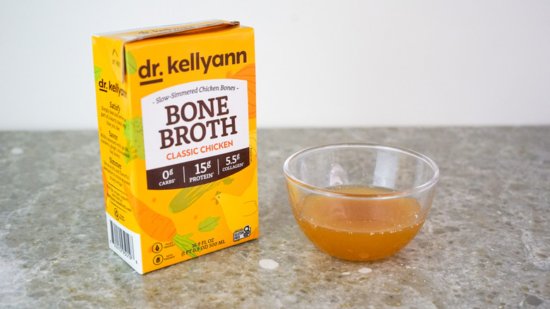 Carton of Dr. Kellyann Bone Broth Classic Chicken next to glass bowl of broth