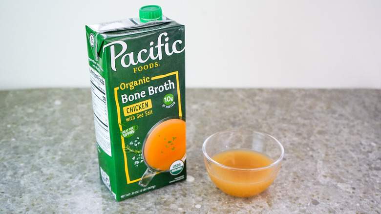 Carton of Pacific Foods Organic Chicken Bone Broth with Sea Salt next to glass bowl of broth