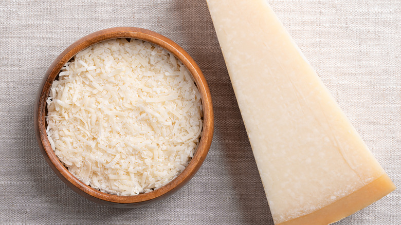 A bowl of grated parmesan cheese next to a wedge of whole parmesan over a woven tablecloth.