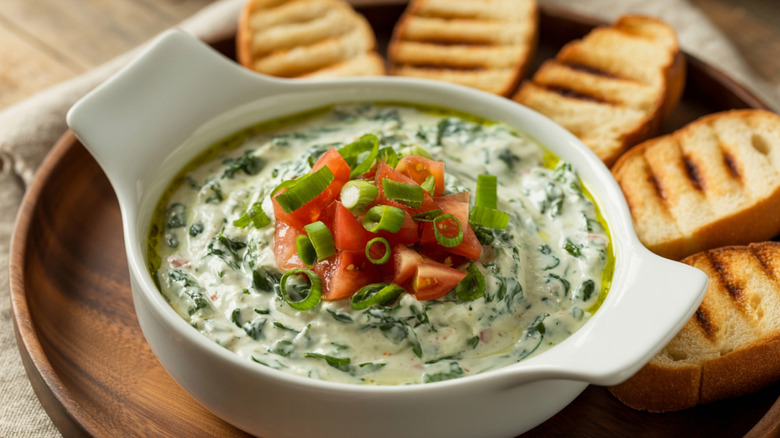 Spinach dip in a white bowl is topped with diced tomatoes and scallions.