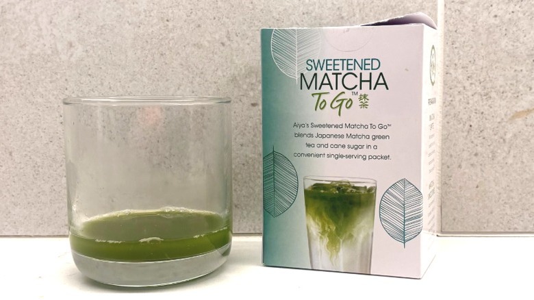 A glass of matcha next to a small white box of matcha powder featuring an image of a matcha drink on a countertop.