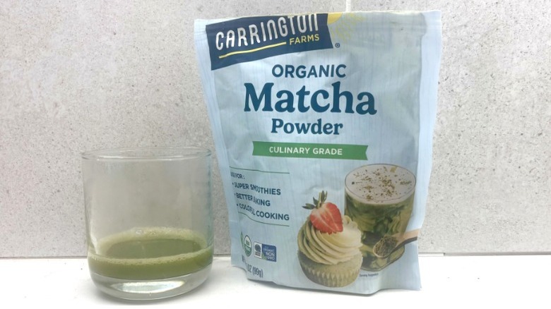 A glass of green matcha next to a sealed blue bag of Carrington Farms matcha powder on a countertop.