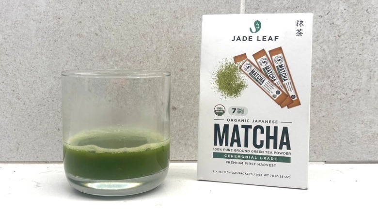 A glass of matcha next to a white box of Jade Leaf matcha powder featuring an image of individual matcha packets on a countertop.